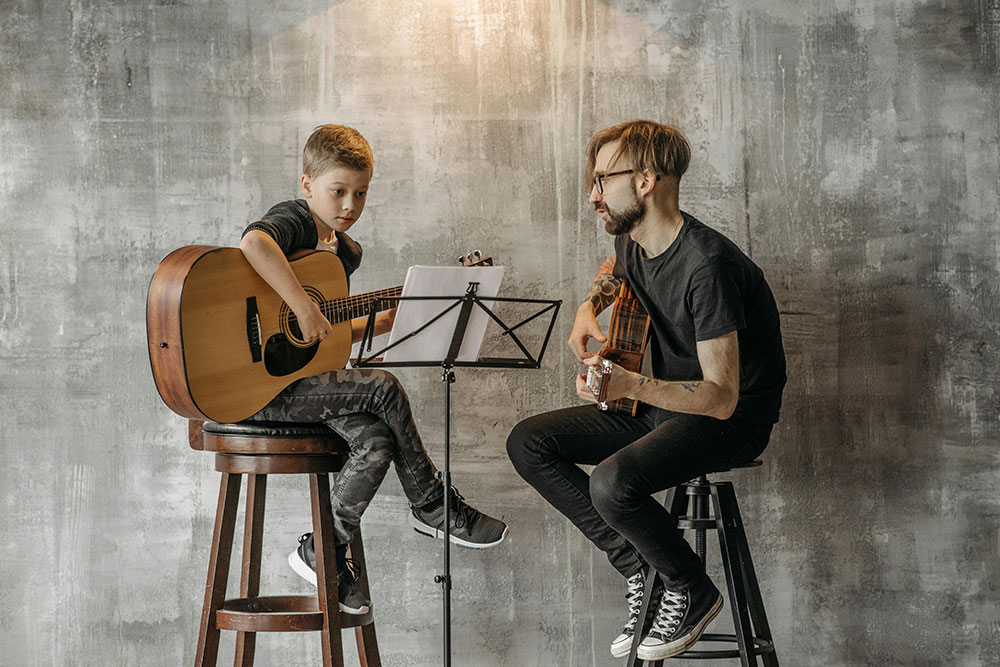 Ein Lehrer und sein Gitarrenschüler spielen ein Stück. Sie sehen dabei auf einen Notenständer.